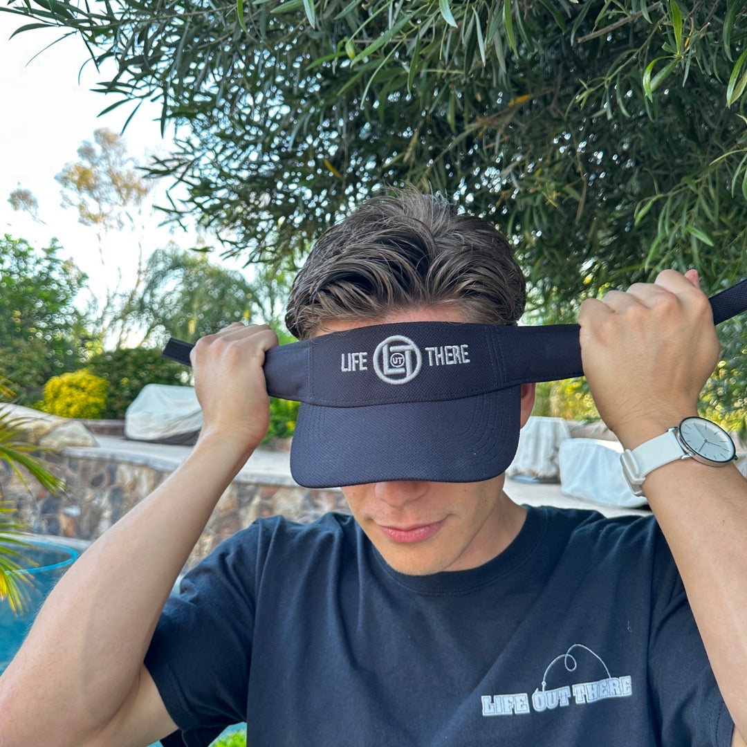 Someone adjusts a black Life Out There - Summer Embroidered Sun Visor under a tree. They are wearing a black t-shirt with the same Life Out There Apparel logo and a watch. The background features greenery and outdoor seating.