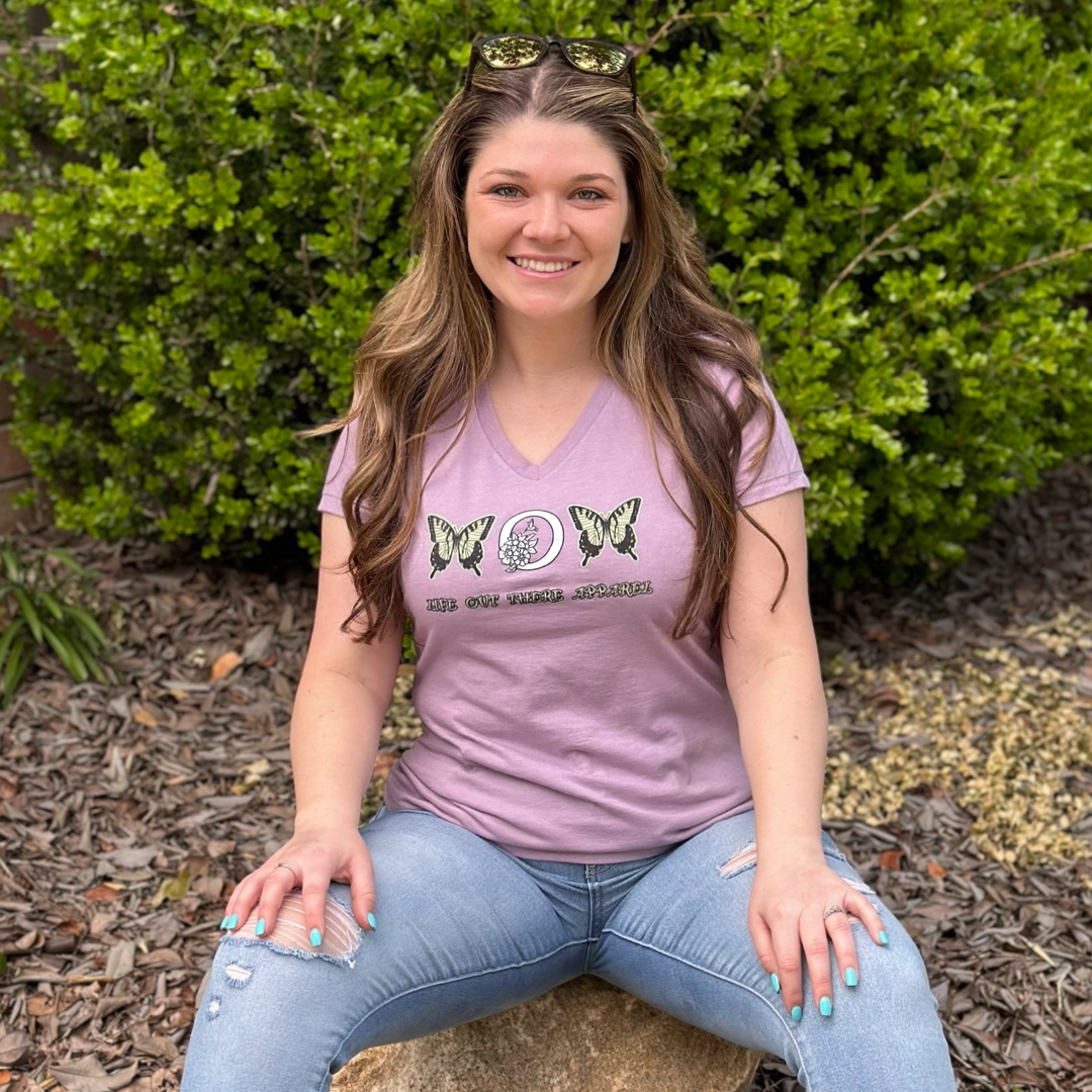 A woman with long hair sits outdoors on a small wooden platform, wearing the Life Out There Apparel's "Love Mom V-Neck T Shirt" in lavender and blue jeans. With sunglasses perched on her head, she exudes tranquility amidst the green bushes surrounding her, creating perfect Mother's Day vibes.