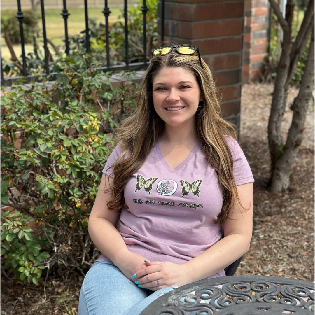 A woman with long hair smiles while sitting at an outdoor table, wearing the Love Mom V-Neck T Shirt in Lavender by Life Out There Apparel, which is beautifully adorned with monarch butterflies. Her jeans complete the casual look against the lush greenery and brick wall behind her, capturing a moment of joy that feels perfect for Mother's Day.
