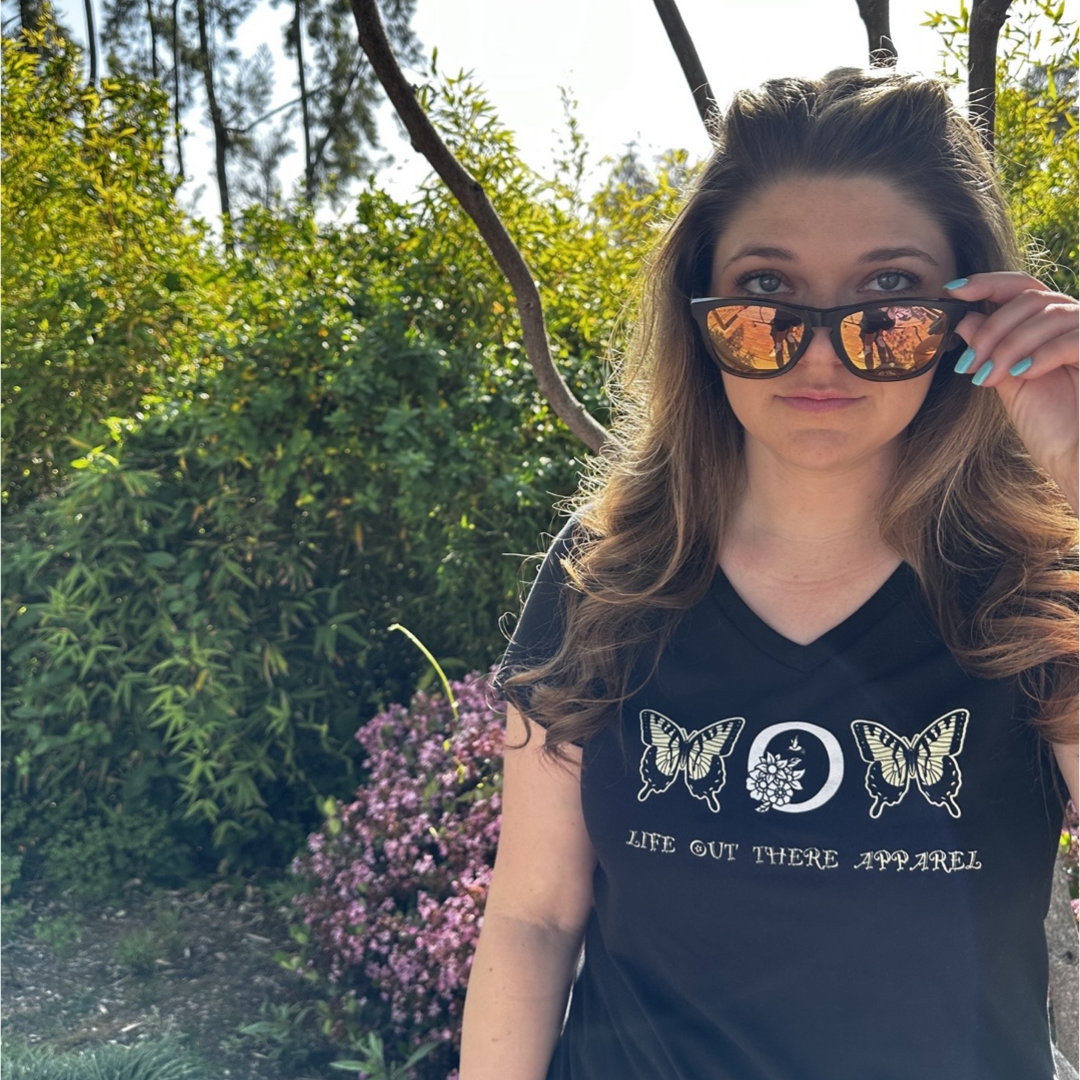 A woman with long brown hair stands outdoors, wearing the Love Mom V-Neck T Shirt in black from Life Out There Apparel, which is adorned with monarch butterfly designs and accessorized with sunglasses. She lifts her sunglasses slightly with her right hand while surrounded by lush green foliage and pink flowers, perhaps celebrating a serene Mother's Day moment.