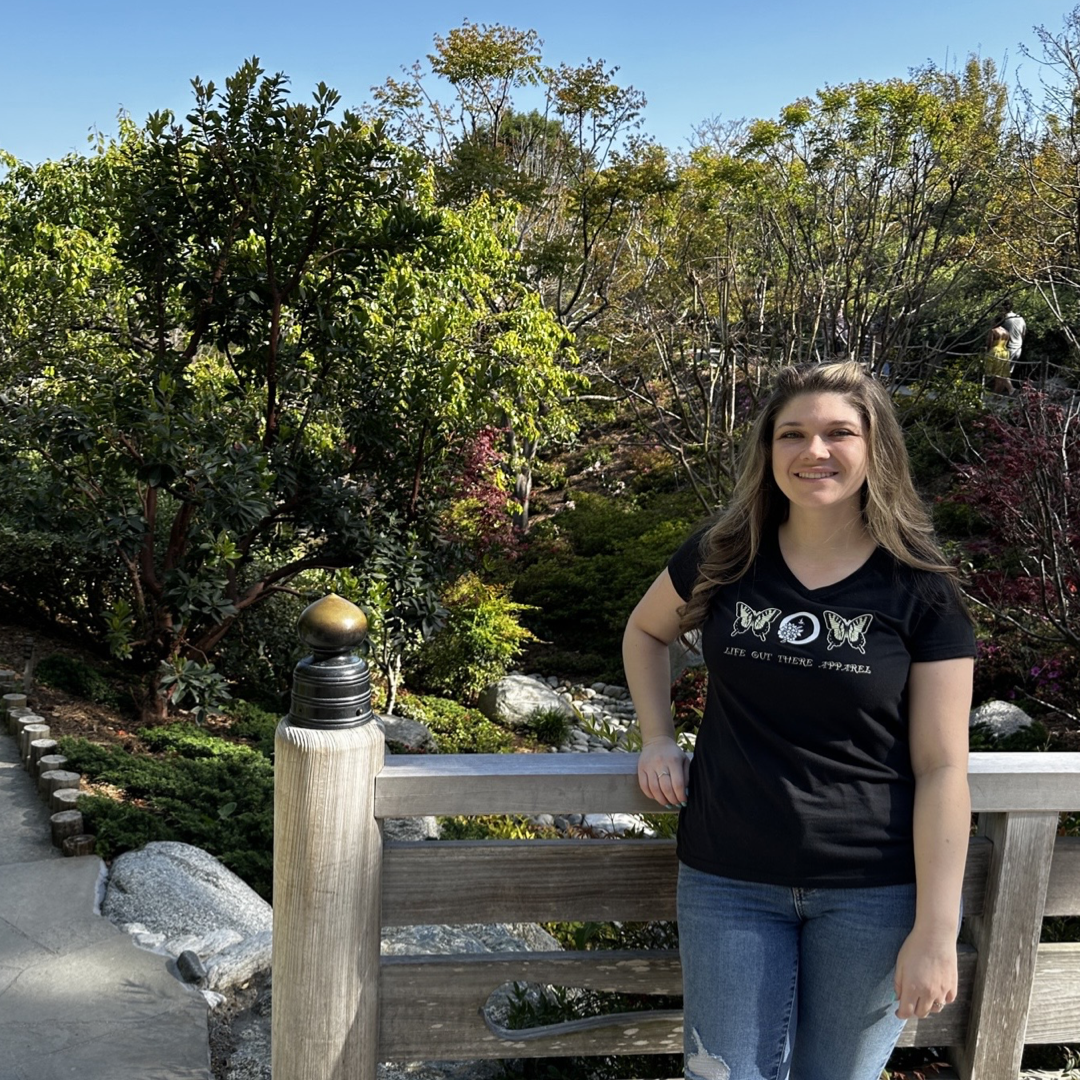 A woman stands on a wooden bridge in a garden filled with lush greenery and vibrant plants, her Life Out There Apparel Love Mom V-Neck T Shirt in black proudly on display. She smiles warmly at the camera as bright sunlight filters through the canopy, mirroring the gentle flutter of a monarch butterfly nearby.