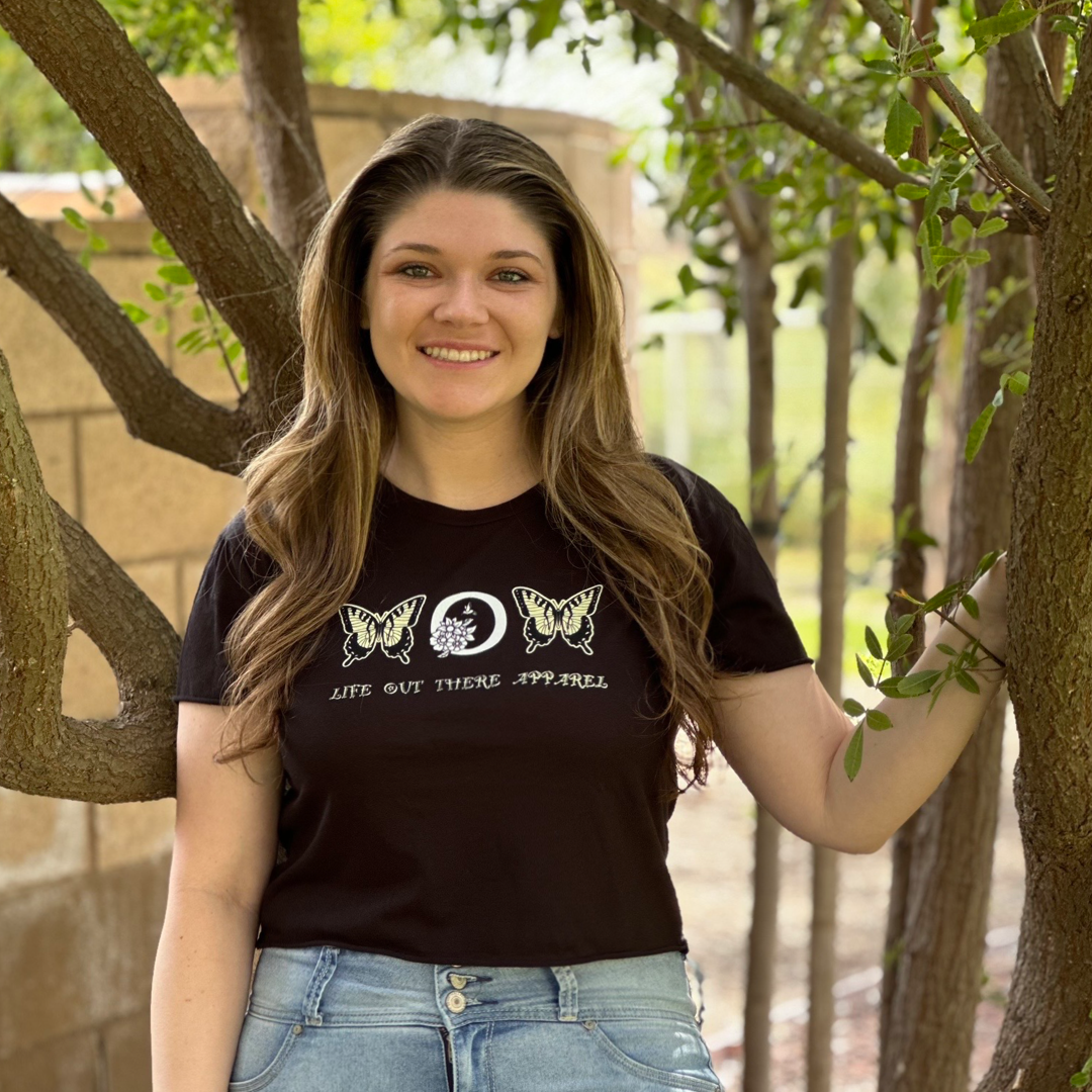 In a lush garden, a woman with long hair smiles, dressed in the "Love Mom Crop T Shirt" from Life Out There Apparel. Her black t-shirt showcases monarch butterfly designs as she gently rests one hand on a tree trunk, celebrating Mother's Day with love.