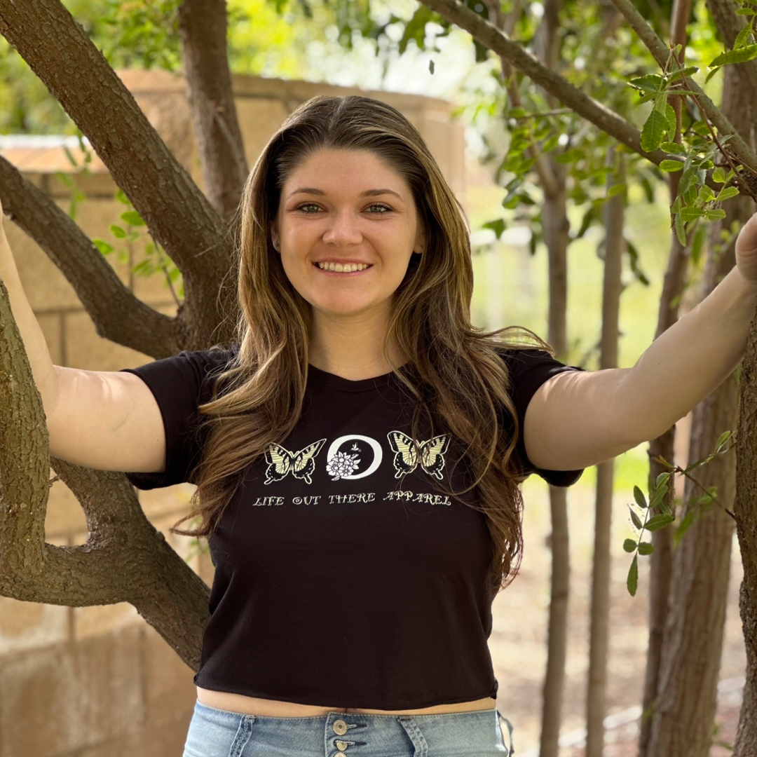 A woman with long hair, wearing the Love Mom Crop T Shirt in black by Life Out There Apparel, stands outdoors. She is smiling and stretching her arms across two tree branches, embodying the spirit of Mother's Day. Lush greenery surrounds her in the background.