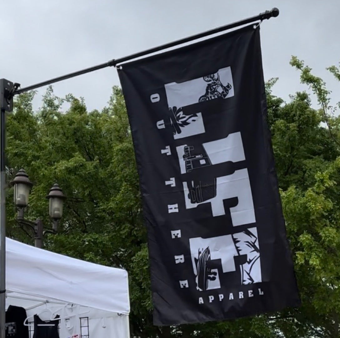 A flag called the OG Flag Black from Life Out There Apparel proudly displays the words "LIFE OUT THERE APPAREL" in bold white letters vertically. Outdoors, it sways against a backdrop of lush green trees and a cloudy sky.