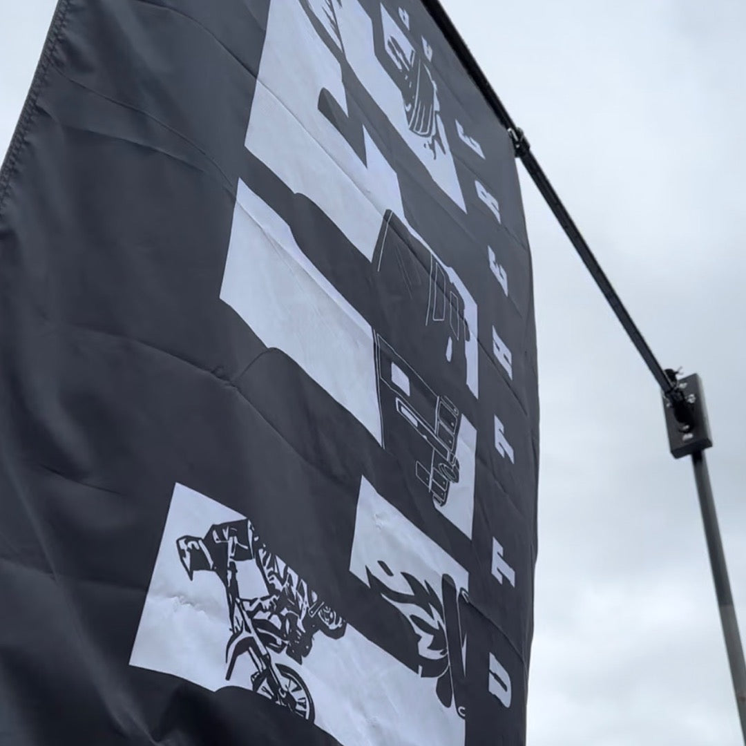 The OG Flag Black from Life Out There Apparel, measuring 3 x 5 and showcasing white abstract designs with mechanical parts and a motorcycle, flutters in the wind against a cloudy sky.