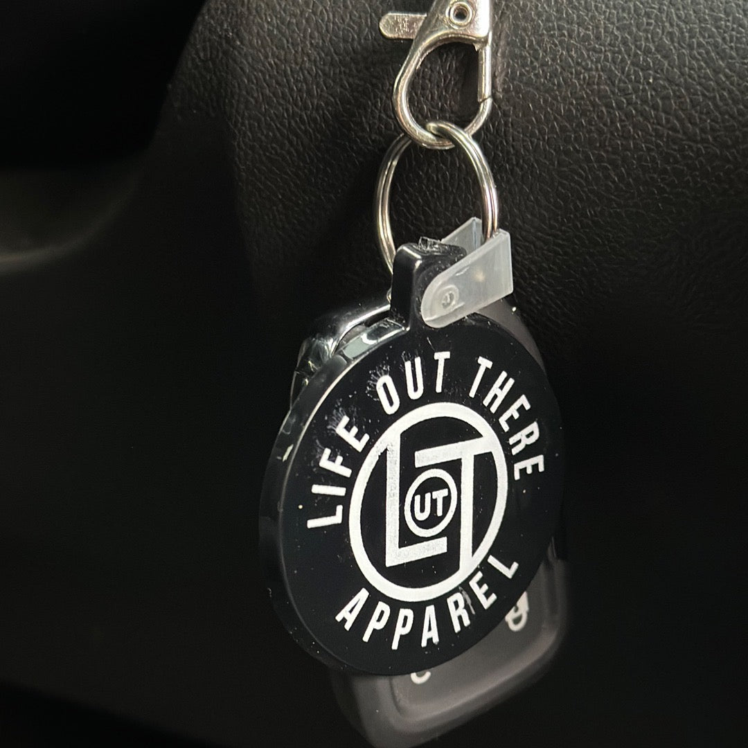 A Life Out There - Black / Round LOT Key Chain from Life Out There Apparel is shown with its round black and white tag featuring the "LIFE OUT THERE APPAREL" text and a central "LOT" logo, hanging against a dark background.