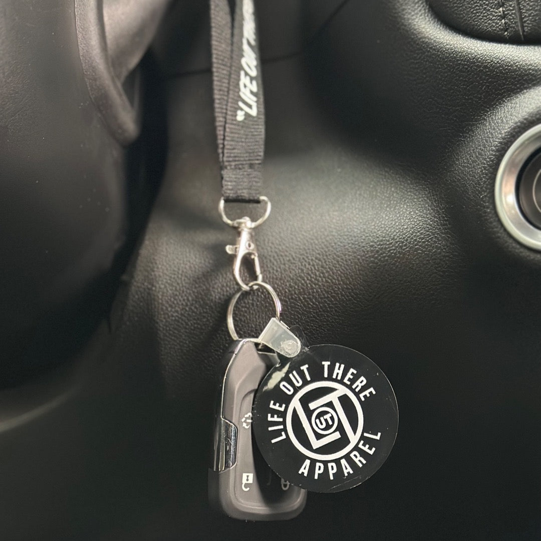 A car key, featuring a Life Out There Apparel's Black Round LOT Key Chain, hangs inside a vehicle against a dark background.