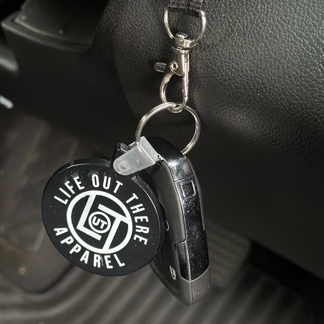 Close-up of a Life Out There - Black / Round LOT Key Chain with the brand name, Life Out There Apparel, visible on the circular tag. The keychain is attached to a car key fob and is hanging in the car's ignition.