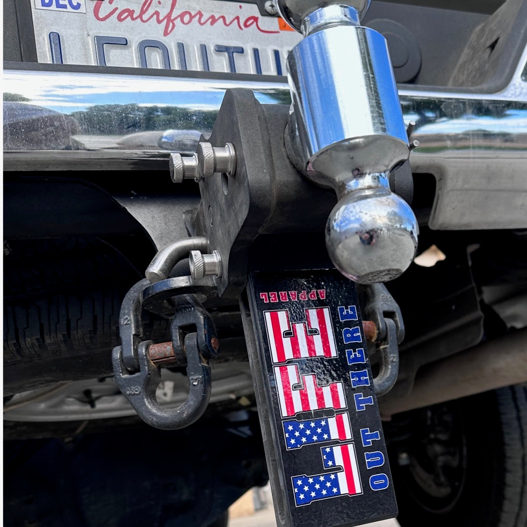 Close-up of a car's rear hitch featuring a hitch cover designed like a red, white, and blue flag with the words LIFE OUT THERE APPAREL. A California license plate is partially visible above it.