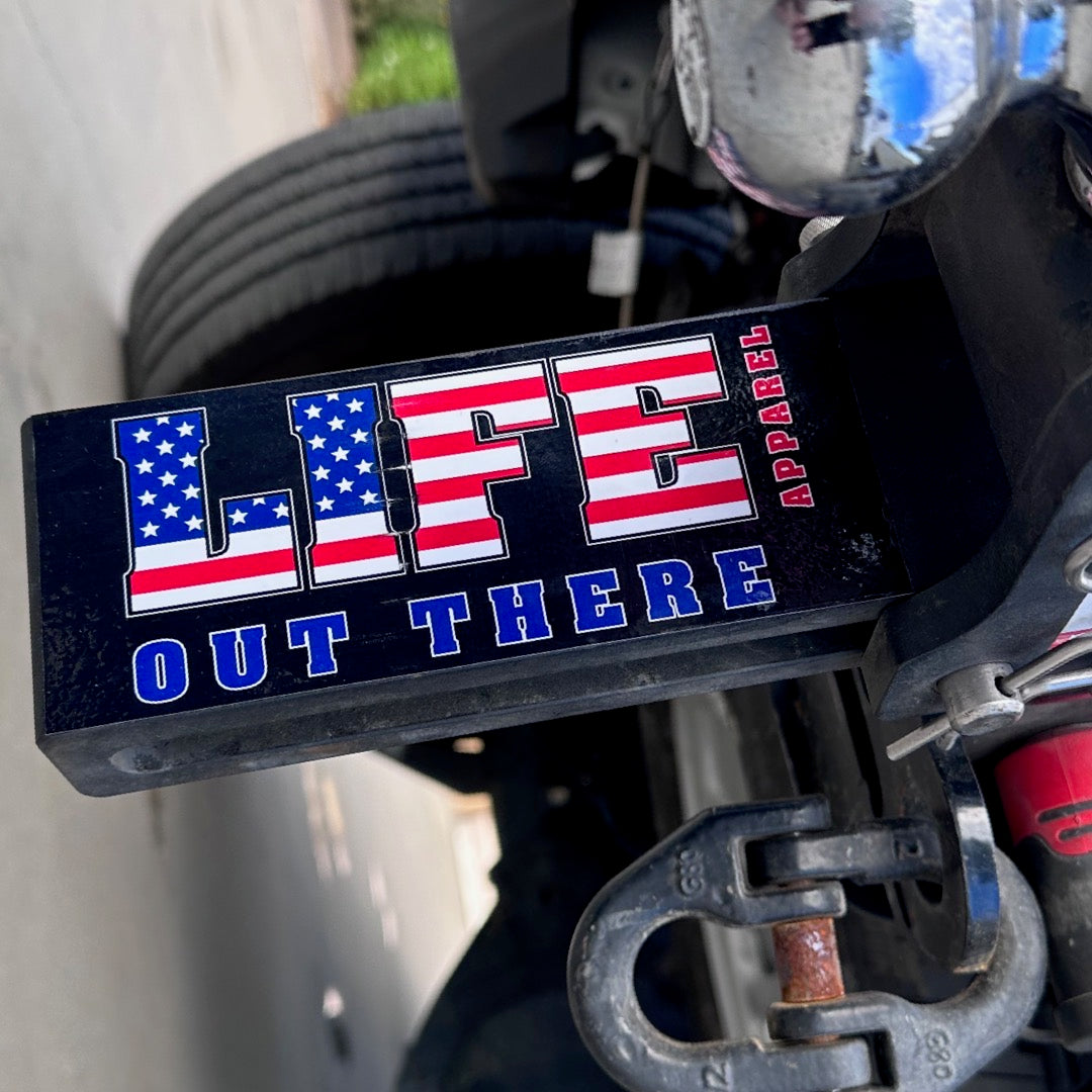 Close-up of a vehicle bumper showcasing the Life Out There - Freedom Sticker by Life Out There Apparel, with "LIFE" designed in the style of an American flag and the words "OUT THERE" in bold. A towing hitch and part of a tire can be seen in the background.