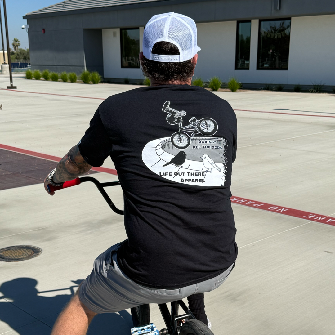 A person in the Unisex - Against All The Odds / The Ride Tee by Life Out There Apparel, gray shorts, and a white cap rides a bicycle on a sunny, paved area.