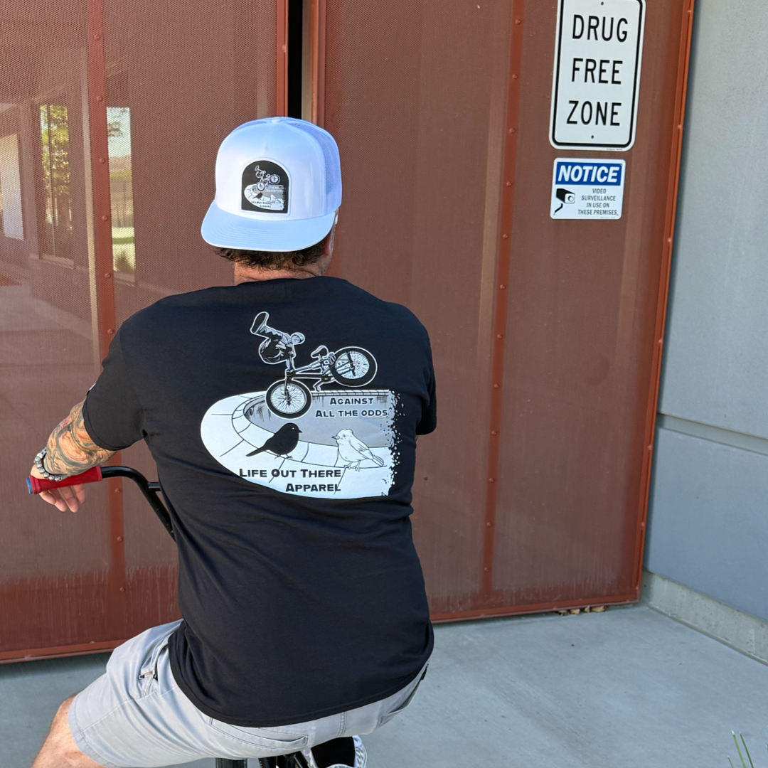 A tattooed man in a Life Out There Apparel Unisex - Against All The Odds / The Ride Tee - Black and white cap sits on a BMX by DRUG FREE ZONE and NOTICE signs, echoing BMX legend John Buultjens spirit.
