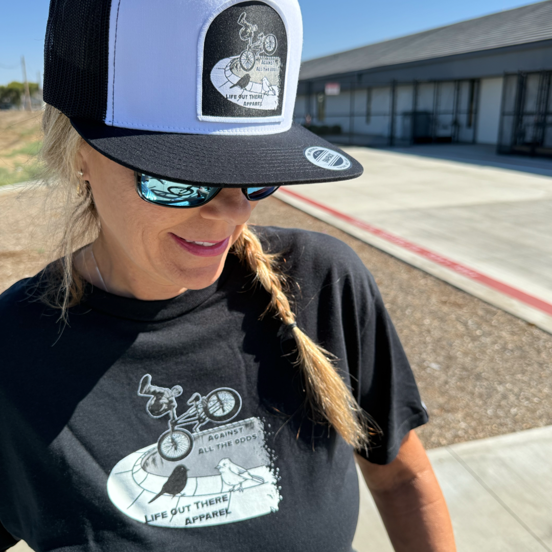 A woman in sunglasses and a braid stands outside wearing a black Against All The Odds / The Ride Tee by Life Out There Apparel and a matching trucker hat, both featuring BMX legend John Buultjens doing a trick.