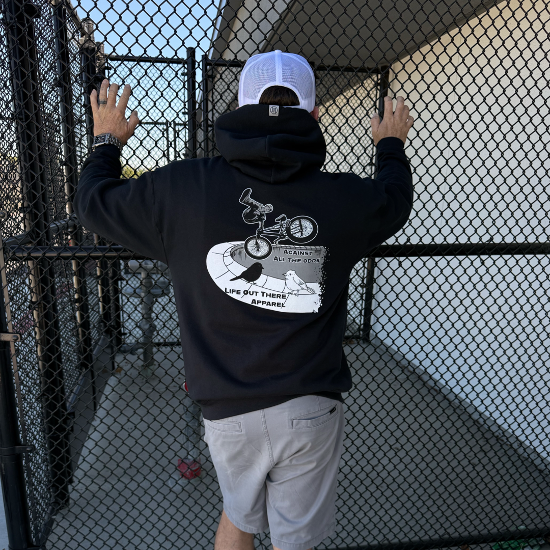 A BMX rider in the Life Out There Apparel Unisex - Against All The Odds / The Ride Hoodie - Black and gray shorts stands at a sunlit chain-link fence, hands gripping it—capturing the spirit of against all the odds.