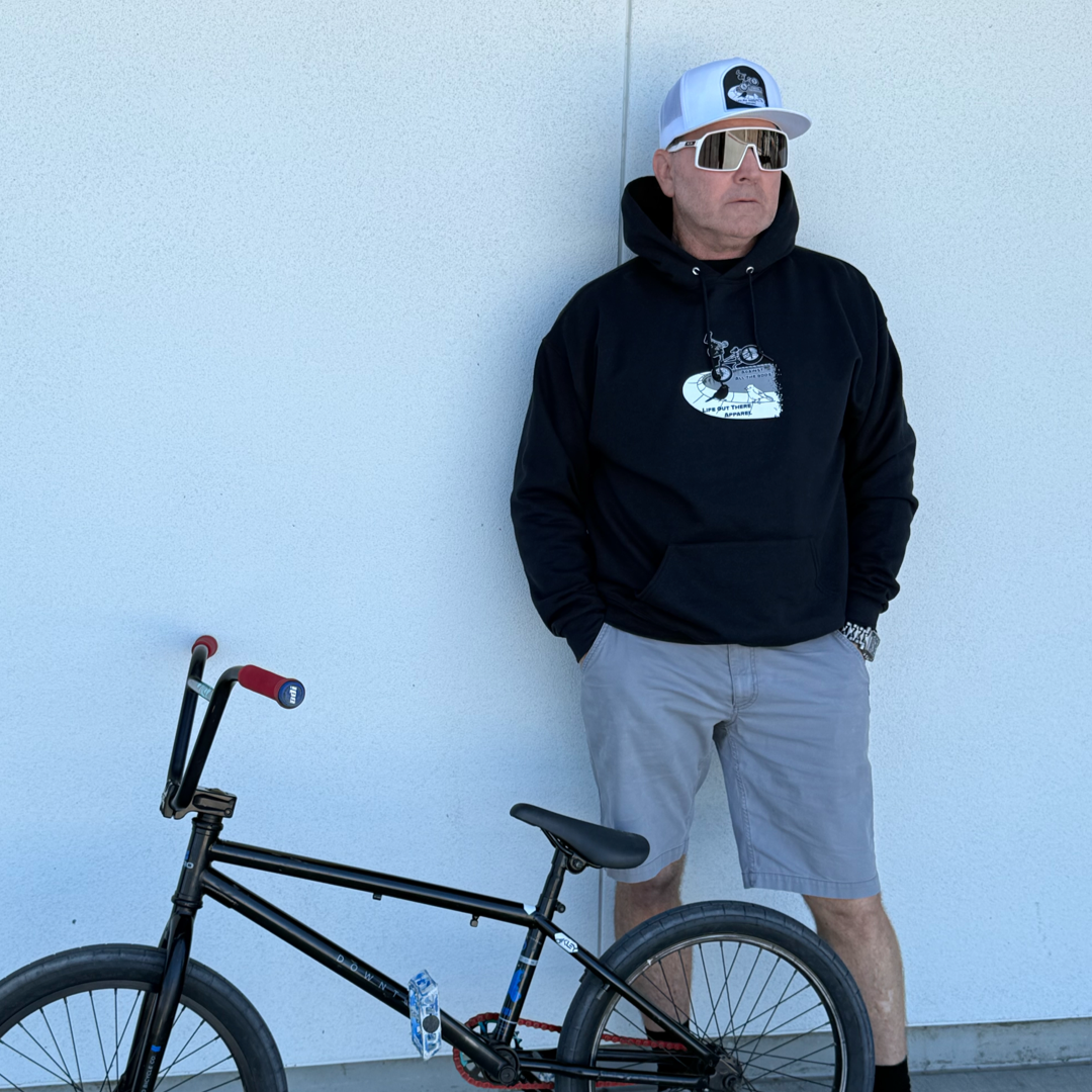 A BMX rider wears the Life Out There Apparel Unisex Against All The Odds / The Ride Hoodie in black, styled with gray shorts, a white cap, and sunglasses. He stands by a white wall with his BMX bike—truly against all the odds.