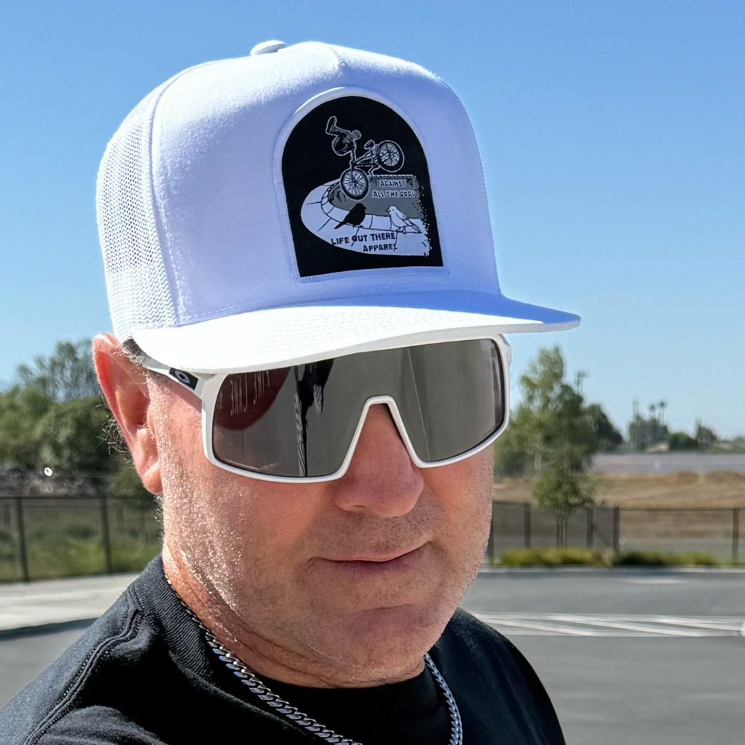 A man wearing the Life Out There Apparel Against All The Odds / The Ride white flat bill snapback trucker hat and white sunglasses stands in a sunny, empty parking lot, channeling BMX rider John Buultjens from THE RIDE movie.