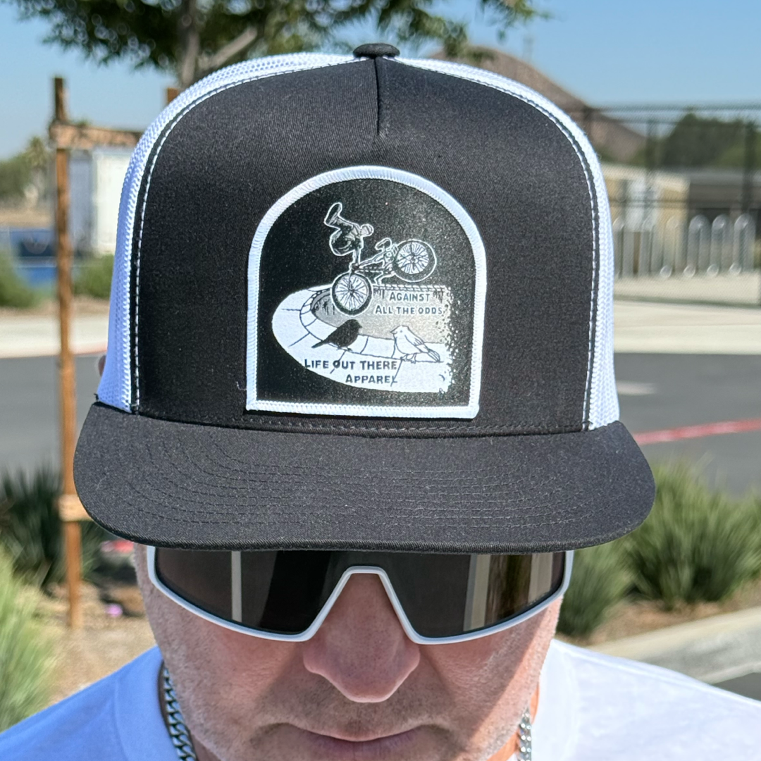 A person wears the Life Out There Apparel Against All The Odds / The Ride Flat Bill Black/White SnapBack Trucker Hat, featuring a BMX rider and outdoor scene, with sunglasses and a blurred background.