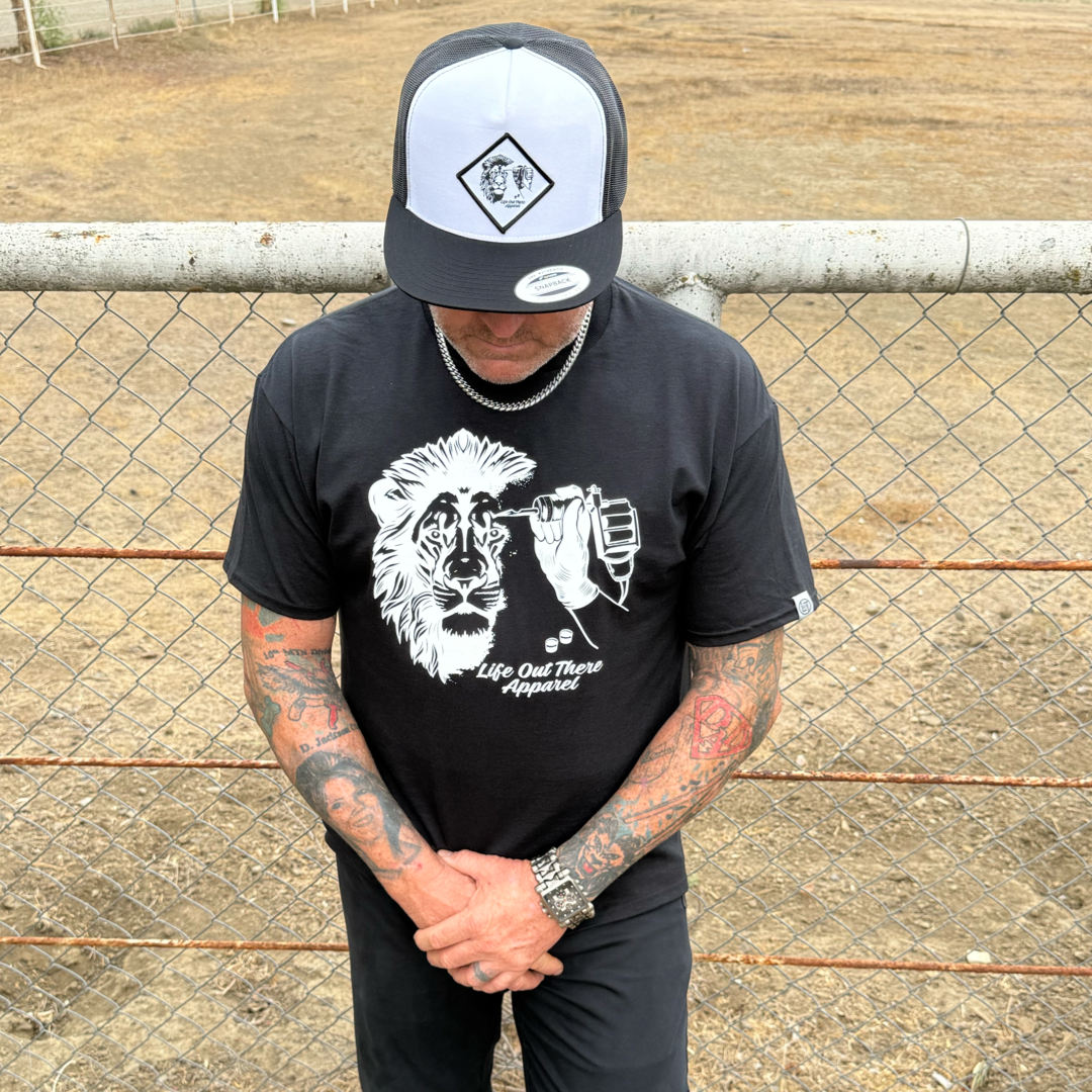 A tattooed man in a black Unisex - Not All There Tee by Life Out There Apparel and a black-and-white hat stands with his head bowed in front of a chain-link fence.
