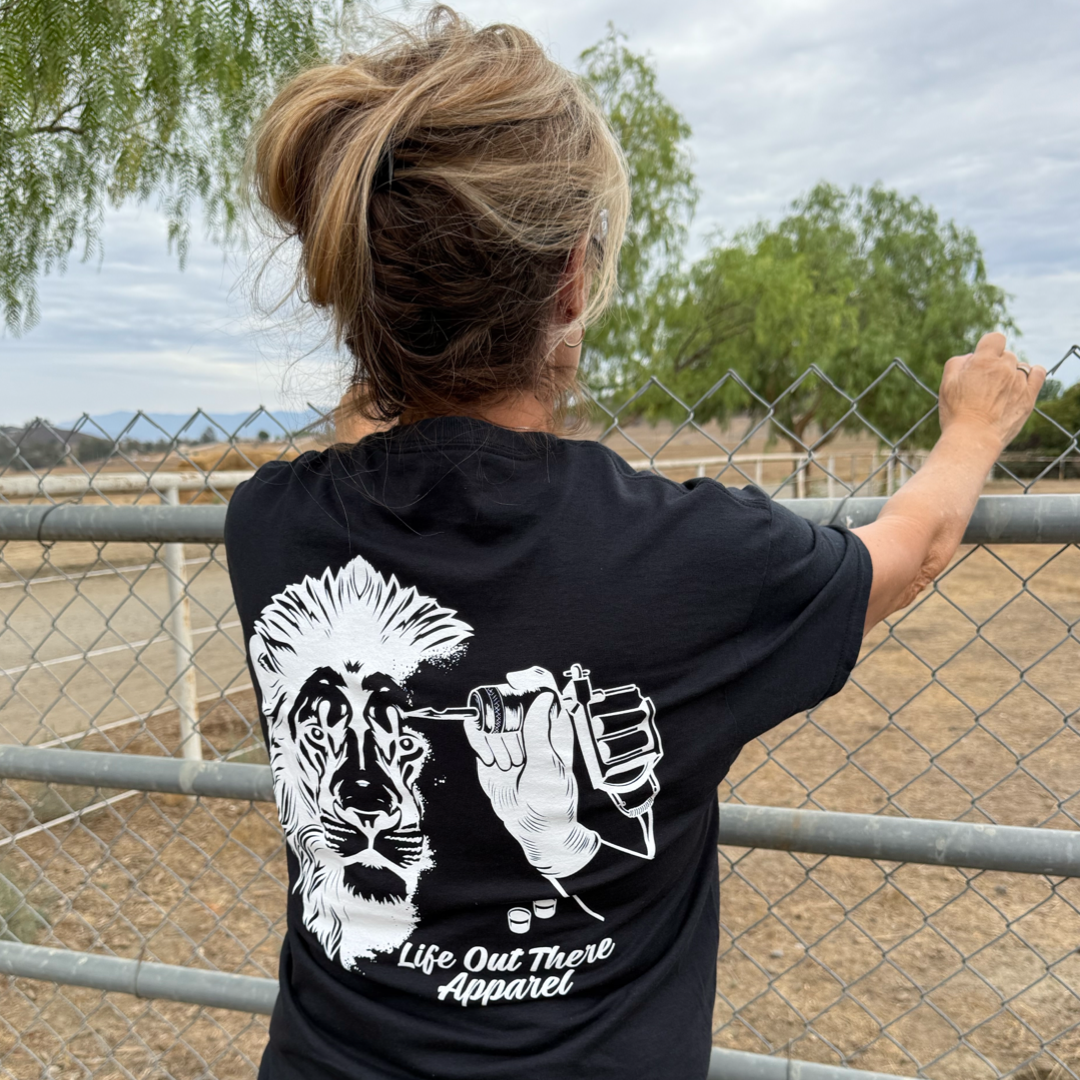 A person stands outdoors, holding a wire fence with trees behind them, wearing the Unisex - Not All There Tee in black by Life Out There Apparel, featuring a lion and tattoo machine graphic on a tagless cotton-polyester blend.