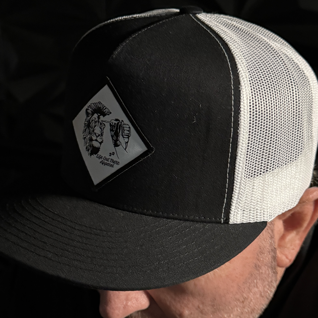 A person wears a Life Out There Apparel Not All There Flat Bill SnapBack Trucker Hat in black and white, featuring a patch with a Spartan helmet, peace sign hand, and the phrase “NOT ALL THERE.”.