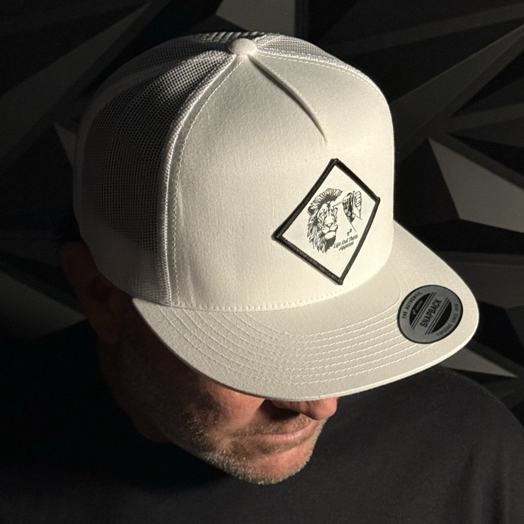 A man wearing the Life Out There Apparel Not All There Flat Bill Snapback Trucker Hat in white, featuring a patch with two hands making the rock on gesture, looks down against a geometric dark background.