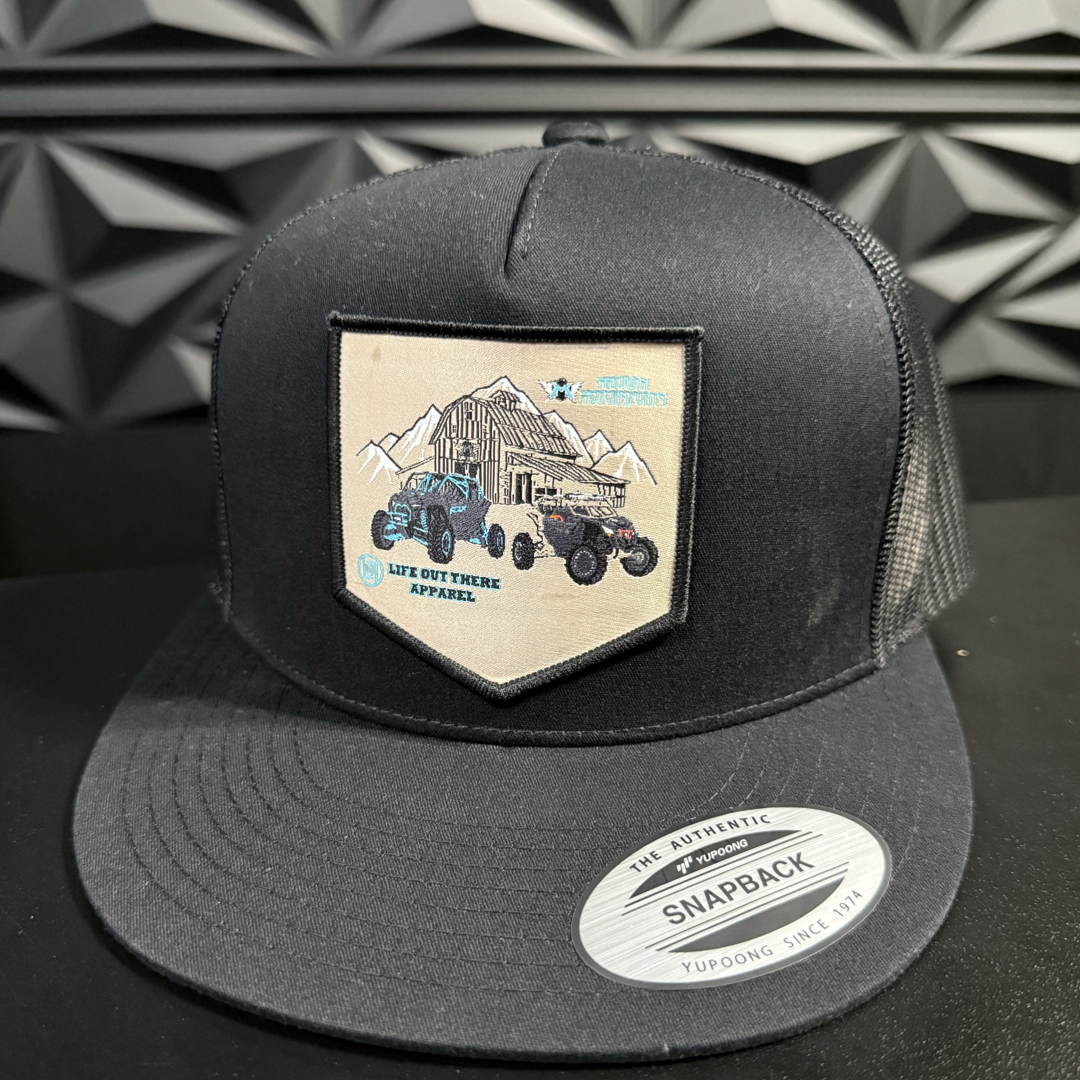 The Side X Side Flat Bill SnapBack Trucker Hat in black by Life Out There Apparel features a patch with off-road vehicles, mountains, and a barn over a geometric-patterned background, echoing an adventurous spirit with Madigan Motorsports.