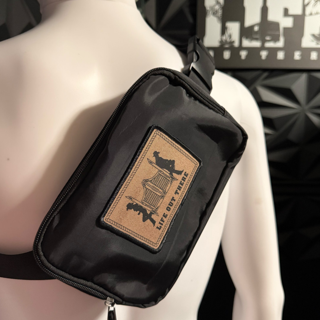 The Yeehaw Fanny Pack / Medium Sized Hand Bag in black by Life Out There Apparel features a brown patch with silhouettes of two people and a tent, plus "LIFE OUT THERE" text, zip pockets, and is shown on a mannequin before a black textured wall.