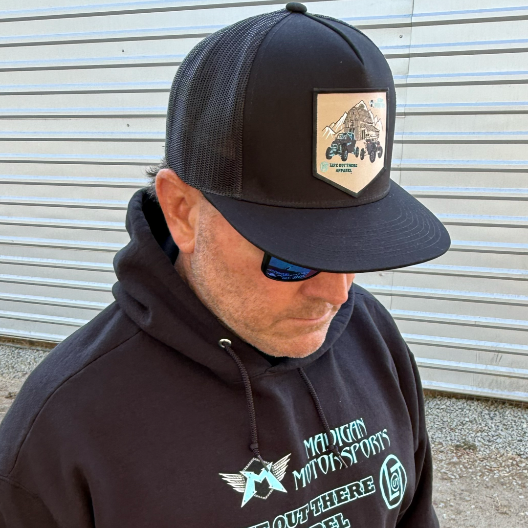 A man in a Life Out There Apparel Side X Side - Flat Bill SnapBack Trucker Hat in black and a “Madigan Motorsports” hoodie stands outside by metal siding, looking down behind blue-reflective sunglasses—ready for adventure.