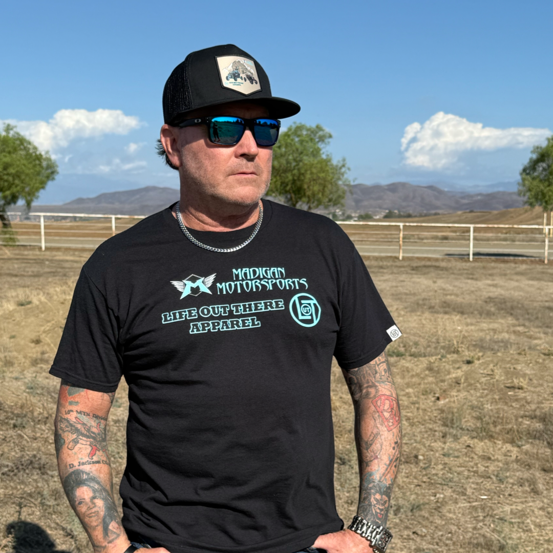 A tattooed man wearing sunglasses, a black cap, and a Life Out There Apparel Unisex Side X Side Tee in black stands outdoors in a dry, open field with trees, mountains, and a partly cloudy sky in the background.