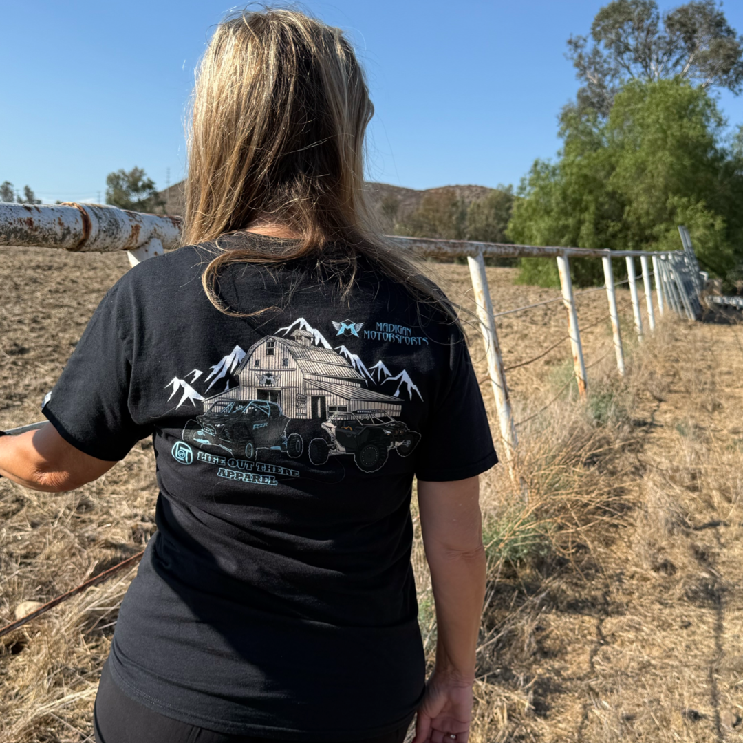 A woman with long blonde hair stands outdoors by a rusty fence, wearing the black Unisex Side X Side Tee from Life Out There Apparel featuring a barn, mountains, and Side X Side vehicles. Dry grass and trees are in the background.