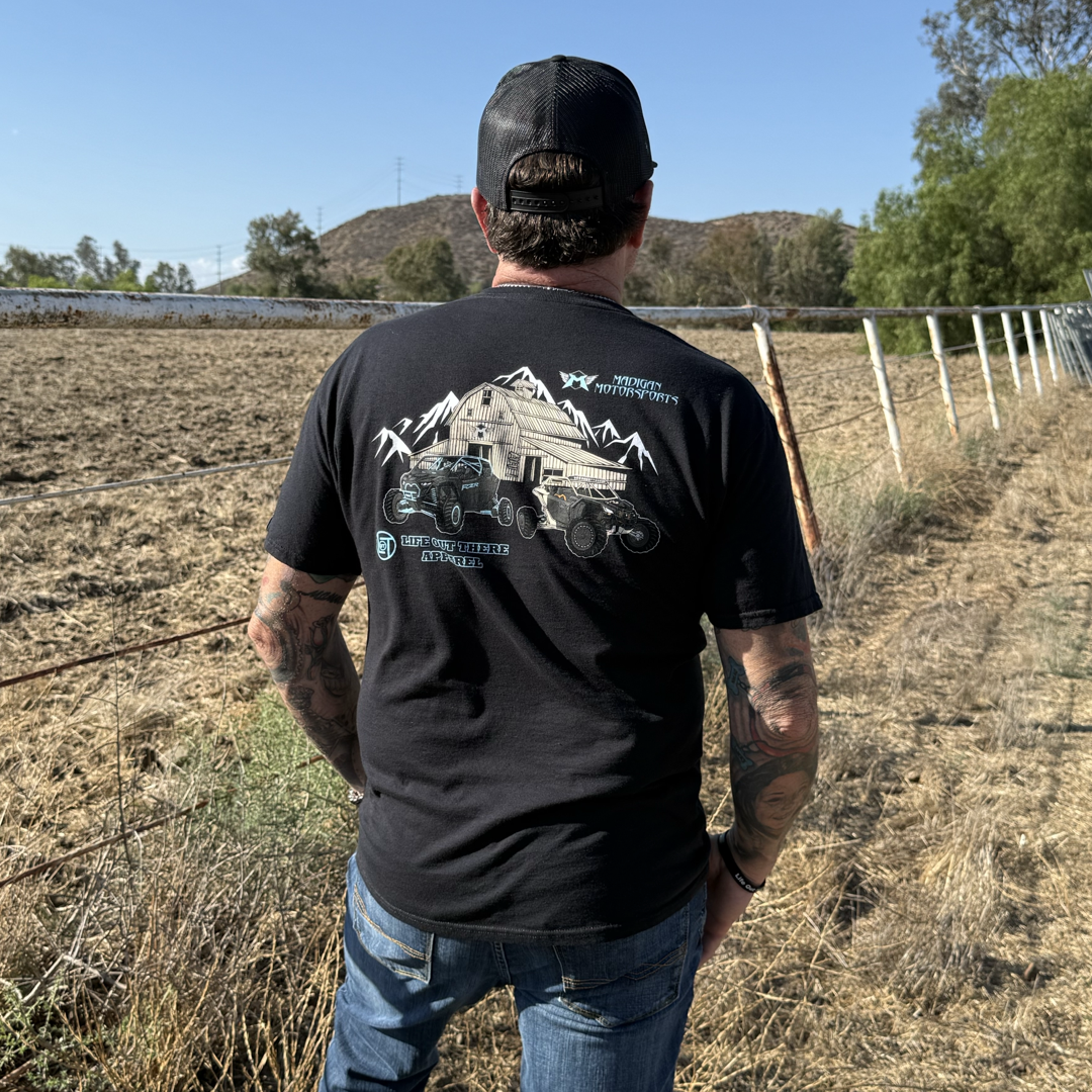 Wearing the Life Out There Apparel Unisex Side X Side Tee in black, a man stands with his back to the camera by a wire fence, looking over a dry field with mountains and trees under a clear blue sky.