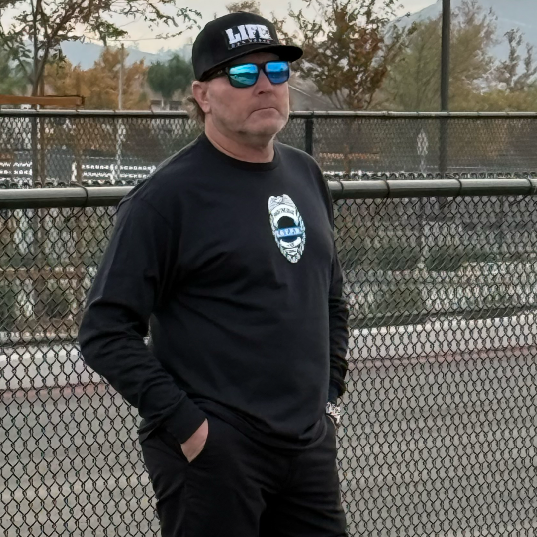 A man sporting a black Back The Blue long-sleeve tee by Life Out There Apparel, a black cap with LIFE, and sunglasses stands with hands in pockets before a chain-link fence, framed by trees and mountains.
