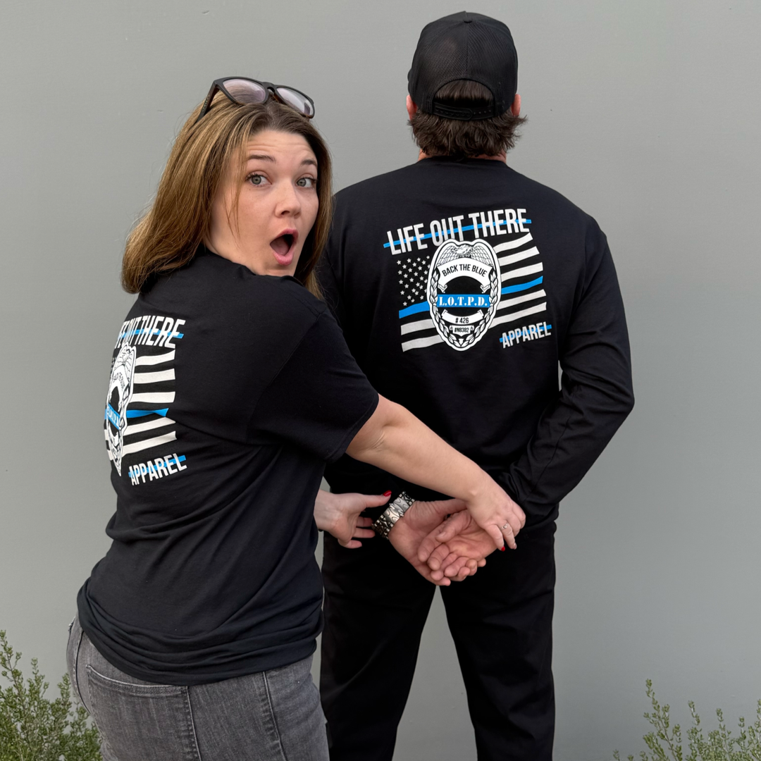 A woman playfully pretends to handcuff a man in a black cap and jeans. Both wear matching Unisex - Back The Blue - Long Sleeve Tee - Black by Life Out There Apparel, standing against a gray wall, embodying the spirit of camaraderie.