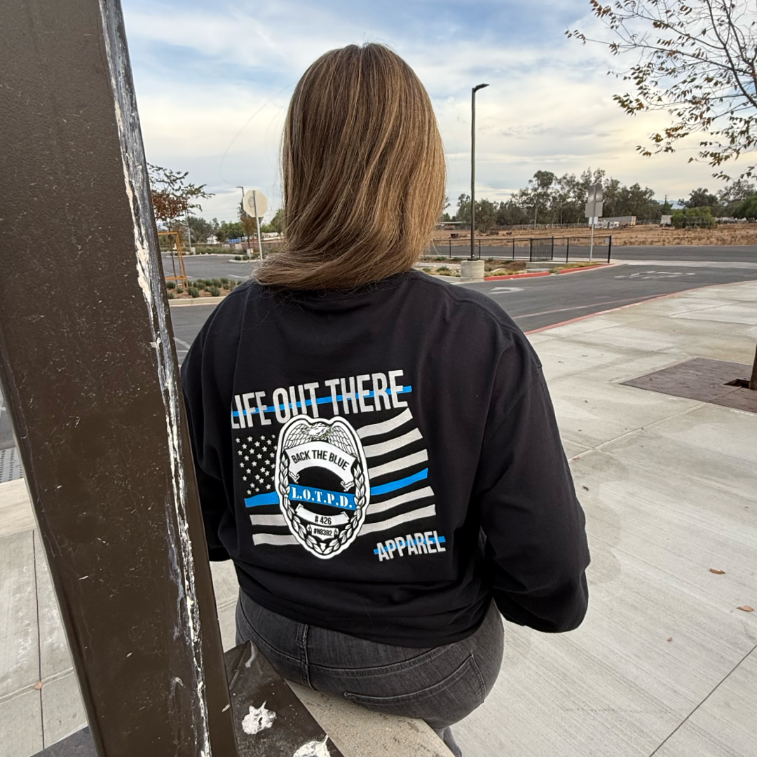 A person sits on a bench wearing the Life Out There Apparel Unisex Back The Blue Long Sleeve Tee in black, featuring the slogan and an emblem. In the background are a street, trees, and a cloudy sky.