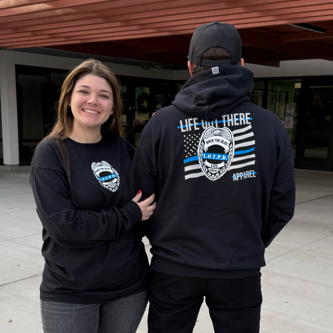 A woman smiles at the camera in a black long-sleeve shirt, while beside her, a man faces away wearing the Unisex - Back The Blue Hoodie by Life Out There Apparel. The hoodie is black with Life Out There and a Sheriffs Department badge design, featuring a blue stripe. Theyre outdoors near a building.