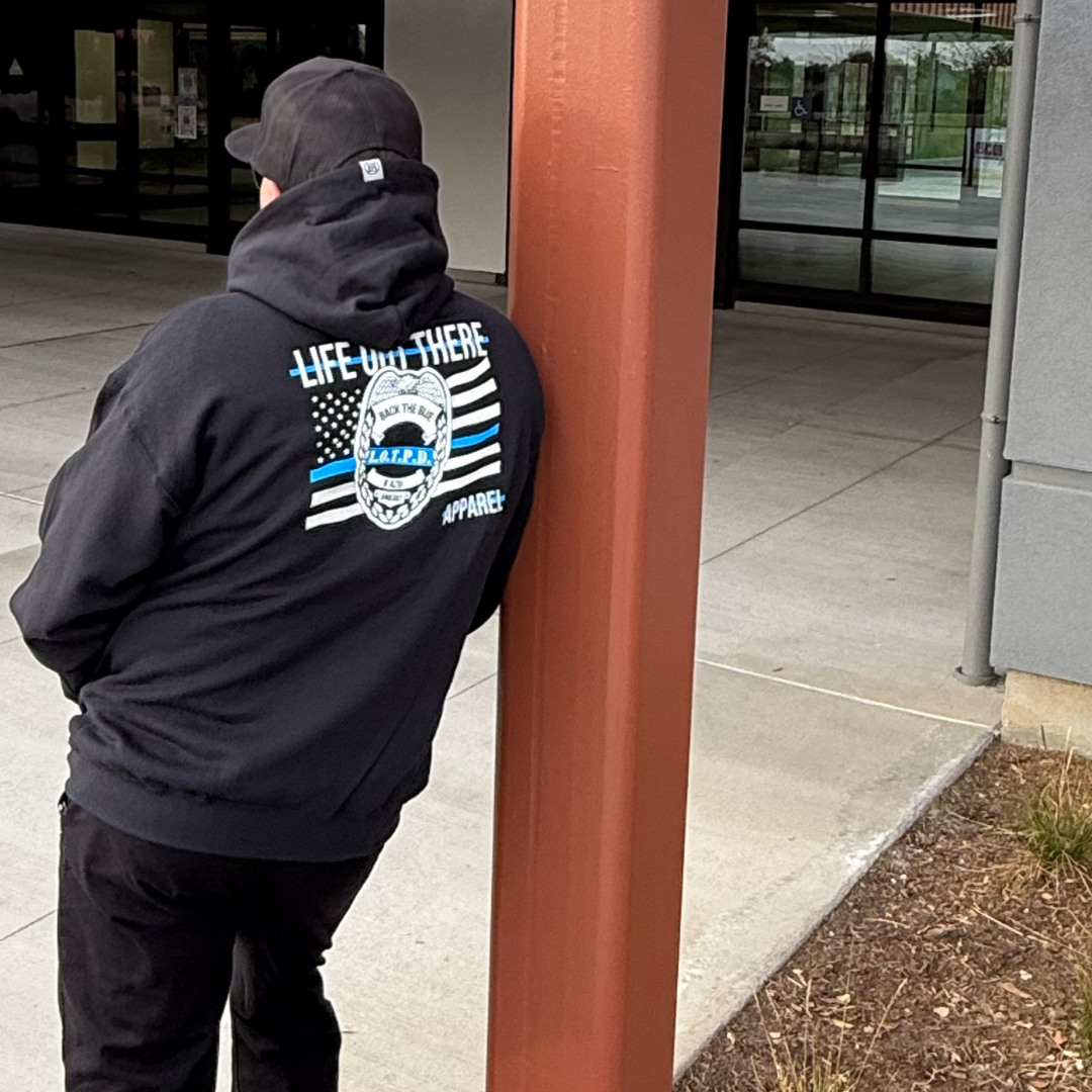 A person, exuding mystery and leaning against a brown pole outside a building, wears the Unisex - Back The Blue Hoodie - Black by Life Out There Apparel, with LIFE OUT THERE and a graphic on the back, giving off a hint of Sheriffs Department vibes while facing away from the camera.