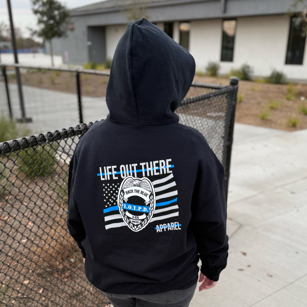 Someone stands by a chain-link fence wearing a black Unisex - Back The Blue Hoodie from Life Out There Apparel. The hoodie features blue and white stripes and a badge on the back. An urban scene with a building and walkway is in the background.