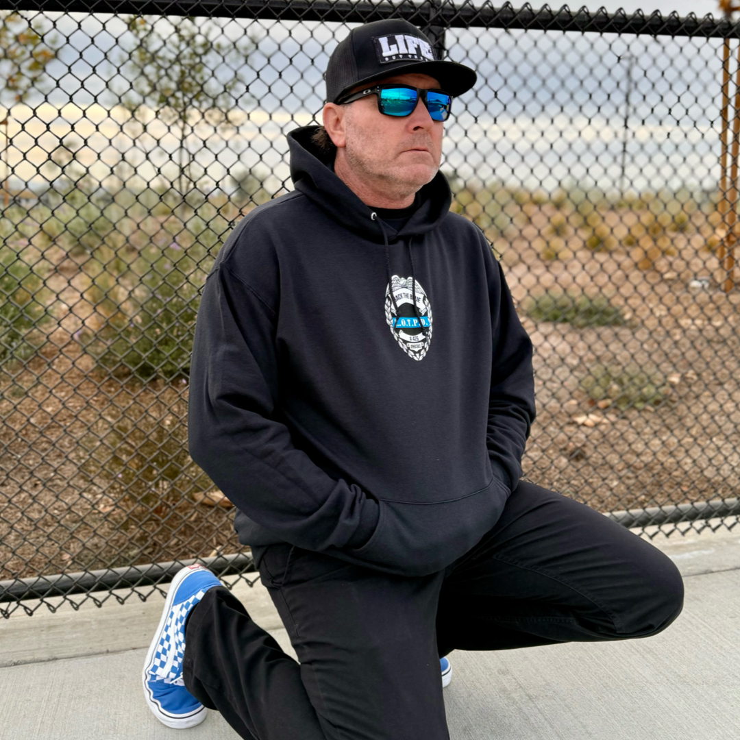 A person kneels on one knee in front of a chain-link fence, donning Life Out There Apparels Unisex - Back The Blue Hoodie in black, a cap, blue sunglasses, and blue patterned sneakers subtly showcasing Back the Blue. The backdrop displays a dry landscape under overcast skies.