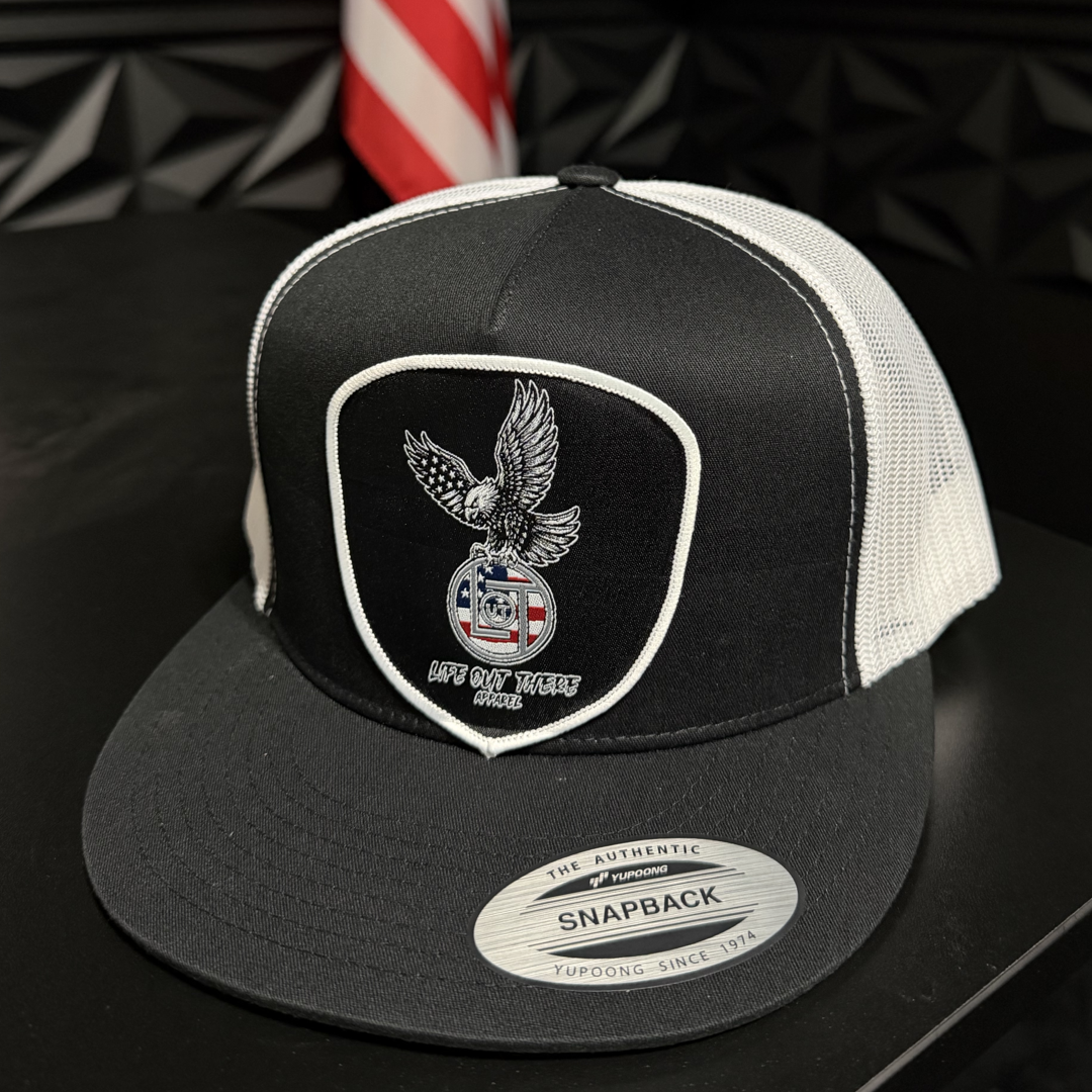 The Flight of Freedom Flat Bill SnapBack Trucker Hat by Life Out There Apparel features a black and white design with an eagle and shield patch, "LIFE OUT THERE APPAREL" and a circular logo. A blurred American flag appears in the background.
