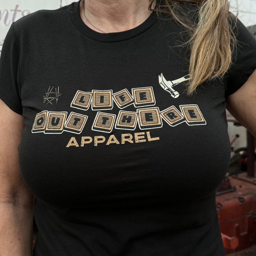 The image features a dad proudly sporting a black t-shirt from Life Out There Apparel, with the brand's name printed in block letters next to a small pickaxe graphic, conveying life lessons through its simple yet meaningful design.