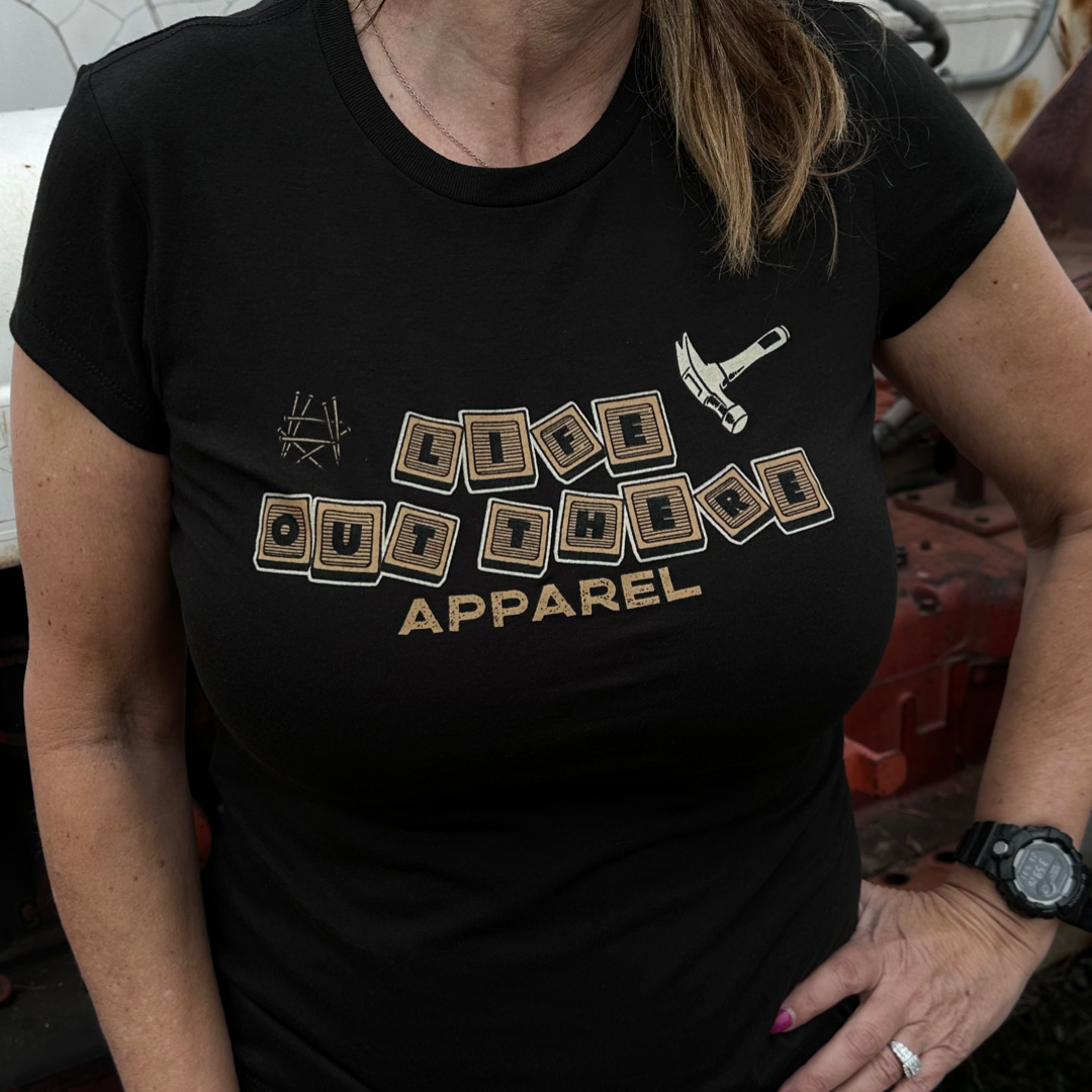 A person is wearing the "Thanks Dad" Women's T-Shirt in black from Life Out There Apparel, featuring "Life Lessons" designed with illustrated wooden blocks and a small hammer. The scene includes a vintage vehicle in the background, with the individual confidently posing with one hand on their hip.