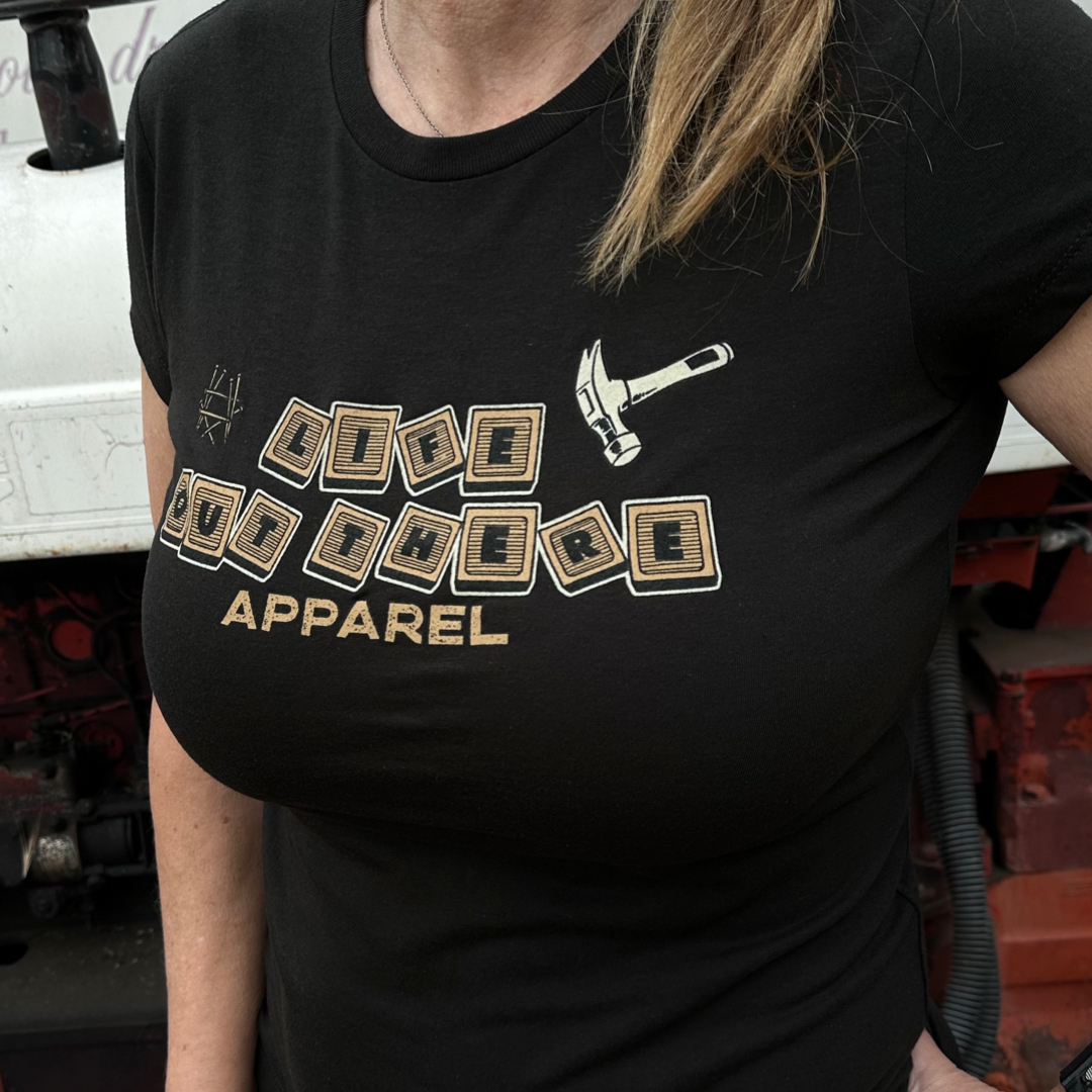 A person is wearing the Thanks Dad Women's T Shirt - Black from Life Out There Apparel, featuring a design with scattered Scrabble tiles and a hammer. The tiles spell out LIFE OUT THERE APPAREL, making it an ideal choice for those embracing life lessons. In the background, part of a vehicle adds rugged charm that Dads would appreciate.