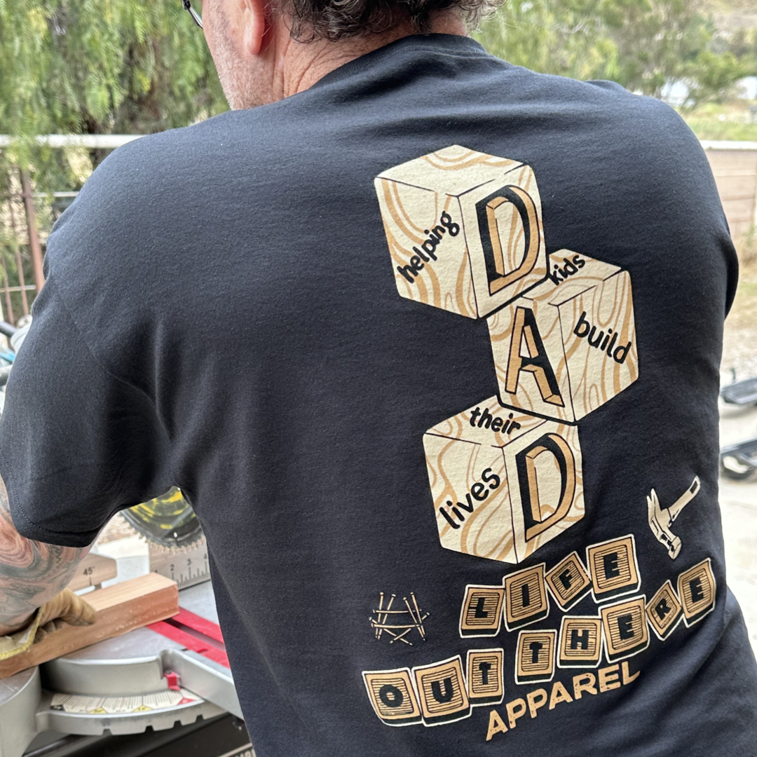 Wearing a black Unisex Thanks Dad T-Shirt from Life Out There Apparel, featuring wooden block designs spelling "DAD" and the phrase "helping kids build their lives," a person skillfully works with woodworking tools outdoors, paying tribute to dads by imparting life lessons through creativity and craftsmanship.