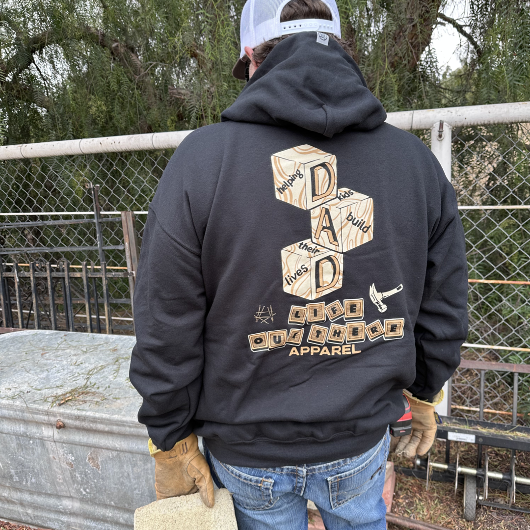 A person is wearing the Unisex Thanks Dad Hoodie - Black by Life Out There Apparel, which features a design of wooden blocks spelling "DAD" and phrases like "builds," "lives," and "shapes life lessons." Standing in front of a metal fence, they're holding a piece of stone while dressed in jeans and gloves. This hoodie celebrates the essence of Dads.