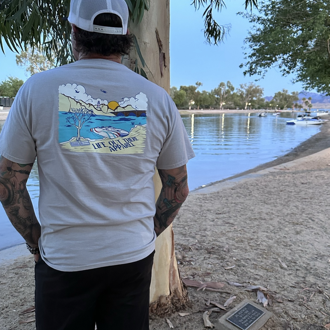 A person wearing a cap and the Unisex The Havasu T Shirt - Silver by Life Out There Apparel stands under a tree by the water, fully embracing the summer vibes. The shirt features the phrase "Life is better at the river" alongside an illustration of a serene Lake Havasu scene with boats docked along the shoreline and trees in the background.