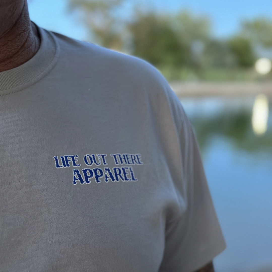 Close-up of a person wearing "The Havasu T-Shirt" in silver by Life Out There Apparel, radiating summer vibes. The background features a blurred outdoor setting reminiscent of the greenery and water at Lake Havasu.