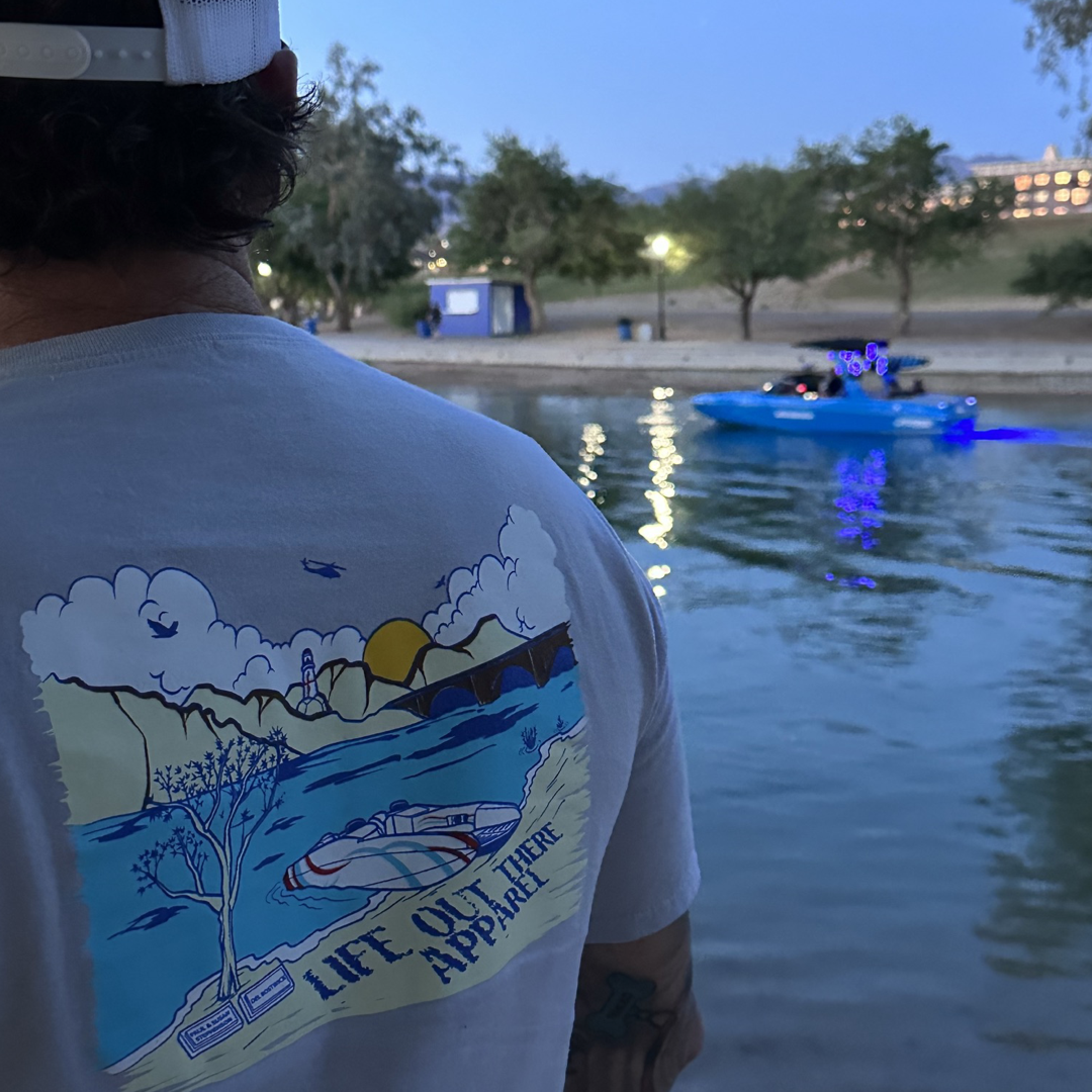 Wearing the Unisex The Havasu T Shirt in Silver by Life Out There Apparel, a person stands by the tranquil Lake Havasu, emanating summer vibes. In the background, a blue boat adorned with string lights drifts on the water, with trees and a building silhouetted against the evening sky.