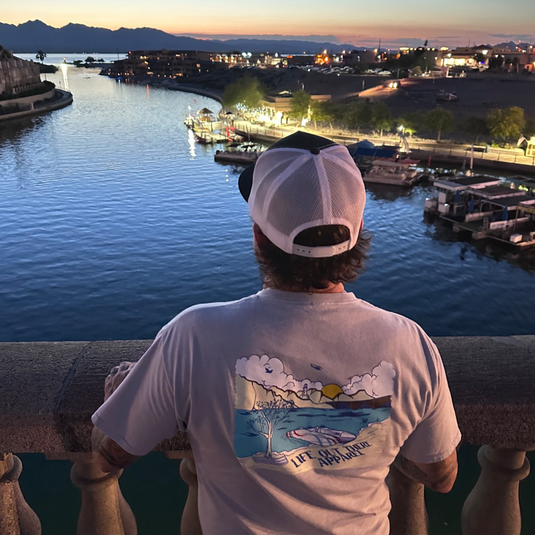 A person wearing a silver unisex "The Havasu" T-Shirt from Life Out There Apparel stands on a bridge, gazing at the river during sunset. The sky blends blue and orange tones, with distant mountains and boats lined along the riverbank, capturing the essence of perfect summer vibes.