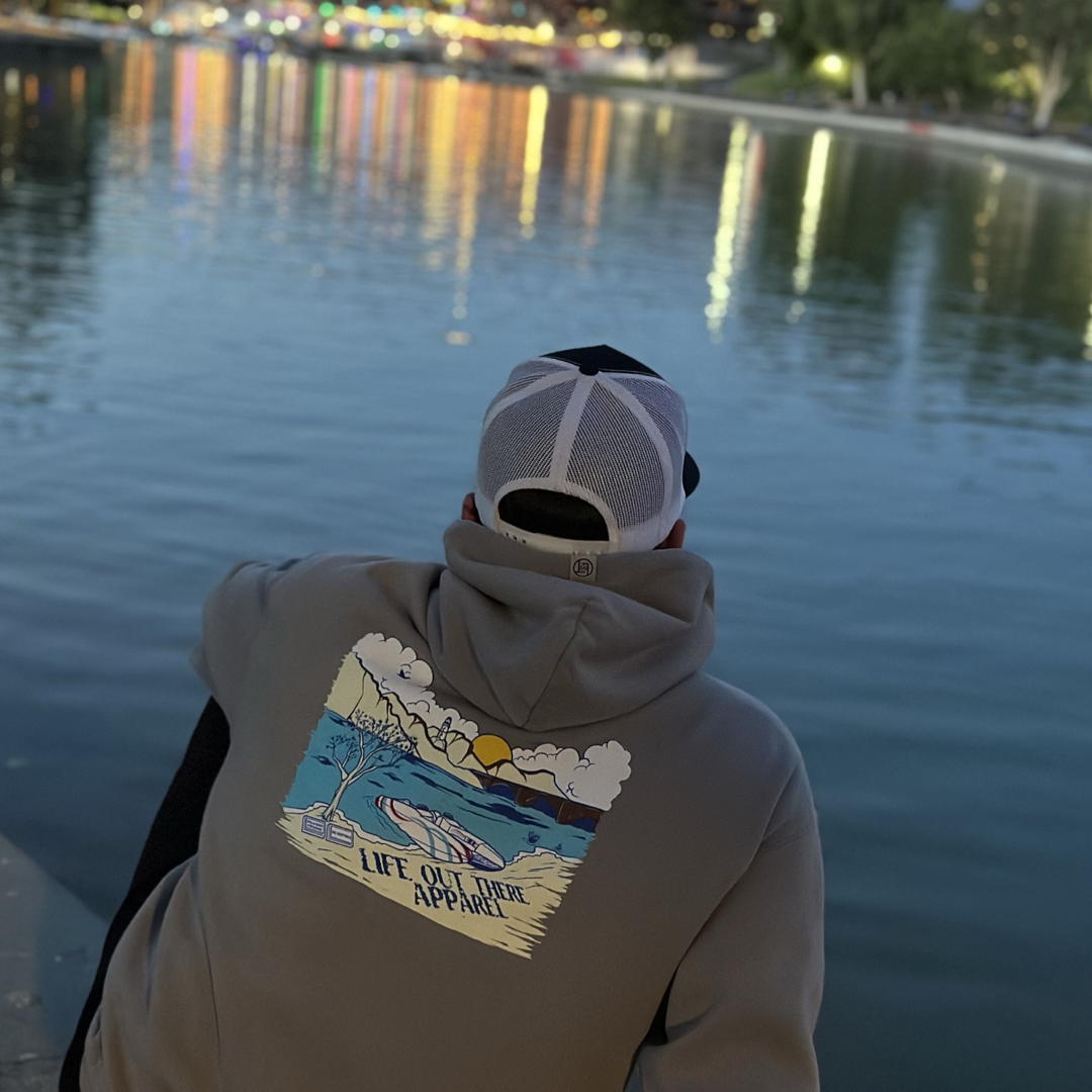 Wearing the Unisex The Havasu Hoodie - Rock by Life Out There Apparel, a person sits by the tranquil waters of Lake Havasu at night. The colorful city lights reflect off the lake, and the back of the hoodie showcases a vibrant graphic that captures Windsor Beach's picturesque landscape.