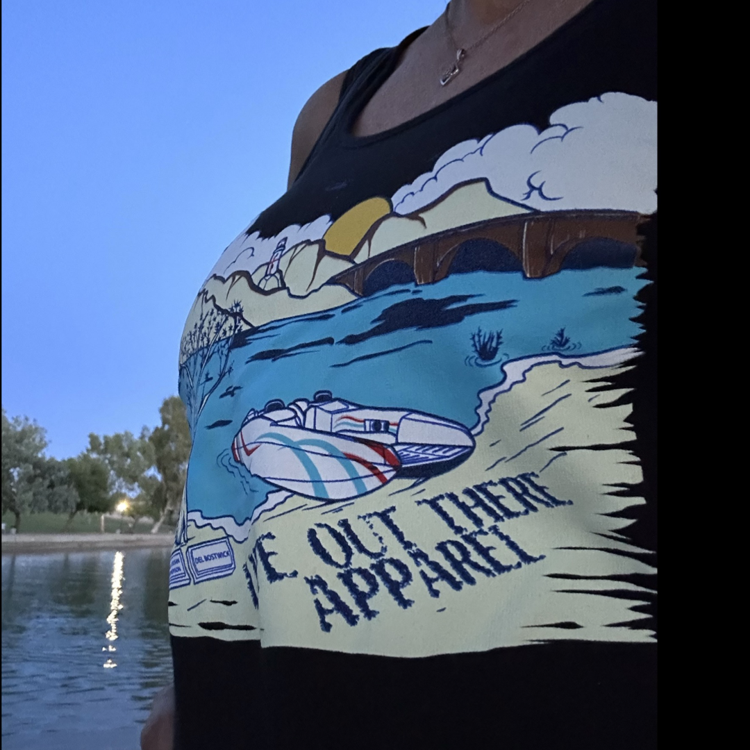 As dusk settles near a body of water, someone is wearing a black tank top showcasing a scenic illustration of Lake Havasu with mountains and a boat, complemented by the text from Life Out There Apparel.